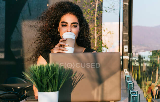 Vue latérale du contenu jeune pigiste ethnique féminine buvant du café et tapant sur le clavier de l'ordinateur portable tout en travaillant sur un projet à distance sur la terrasse du café — Photo de stock