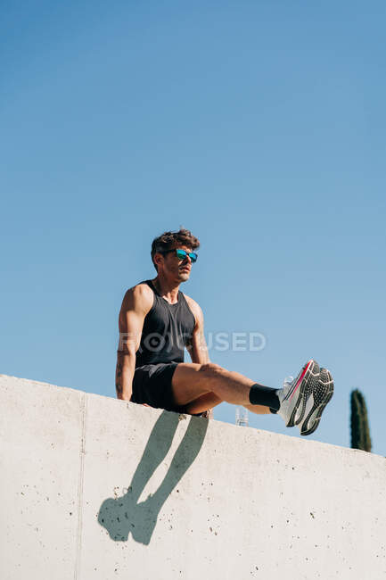 Снизу спортсмен в спортивной одежде и солнечных очках тренируется на заборе под безоблачным небом под солнечным светом — стоковое фото