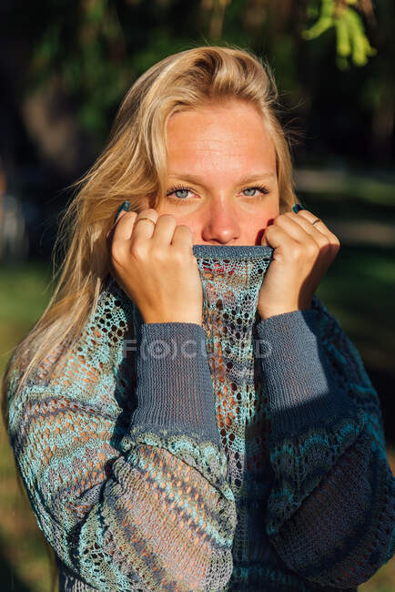 Délicate femelle aux cheveux blonds et en pull tricoté regardant la caméra dans le parc au coucher du soleil — Photo de stock