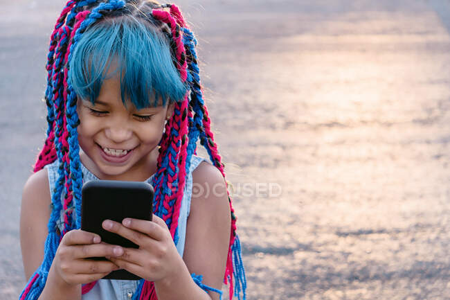 Cheerful Mexican child with colorful braids surfing internet on cellphone in evening on blurred background — Stock Photo
