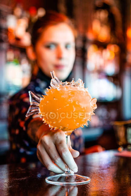Unscharfe Barkeeperin mit alkoholorangefarbenem Cocktail in fischförmigem Glas auf Theke in Bar gestellt — Stockfoto