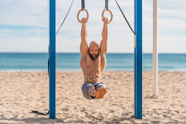 Мужчина без рубашки с бородой висит на гимнастических кольцах с поднятыми ногами, упорно тренируясь на песчаном пляже, глядя вниз — стоковое фото