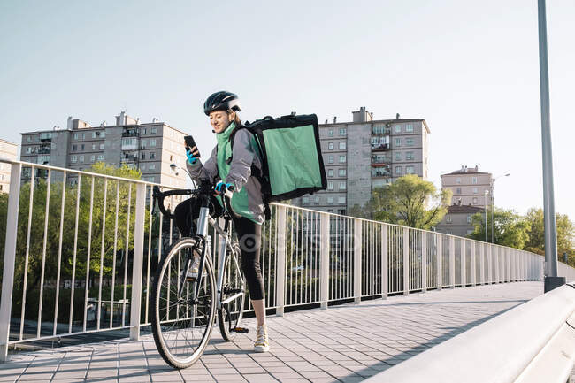 Corpo inteiro jovem mensageiro feminino em uniforme e capacete sentado na bicicleta e navegação mapa GPS no celular na ponte na cidade — Fotografia de Stock