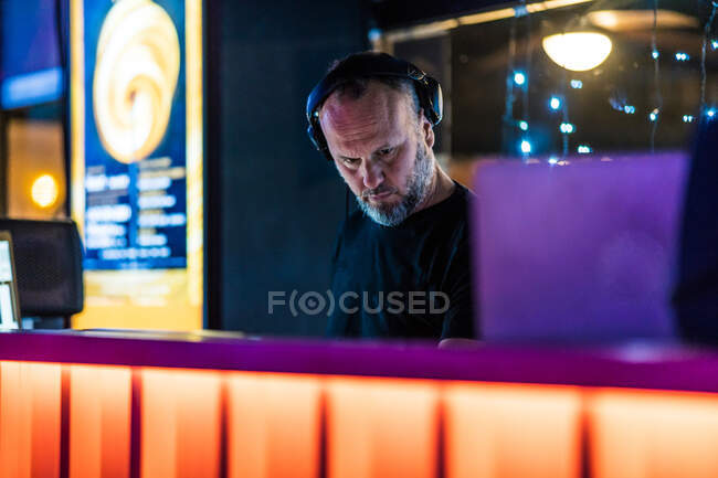 Bajo ángulo de hombre serio de mediana edad en auriculares inalámbricos que se concentran y juegan controlador DJ mientras se realiza en concierto en el vibrante mostrador naranja en el escenario en la discoteca moderna - foto de stock