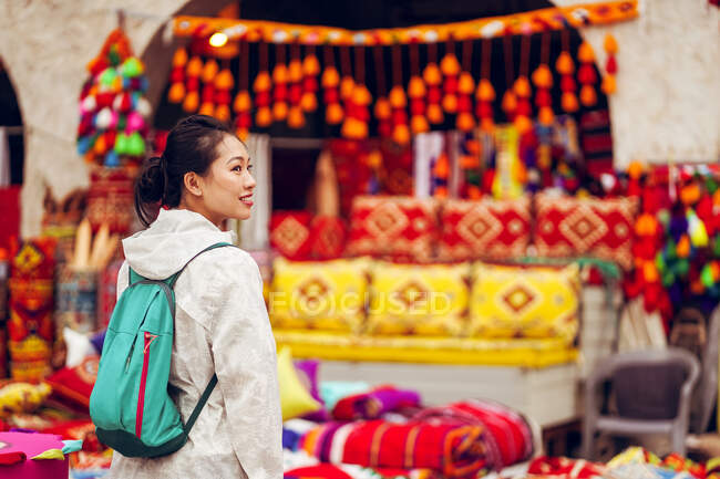 Zufriedene asiatische Touristin mit Rucksack lächelt weg, während sie auf Basar in Katar vor bunten traditionellen orientalischen Kleidern und Souvenirs steht — Stockfoto