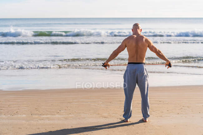 Vista posterior de un hombre brutal irreconocible sin camisa haciendo moscas laterales laterales con entrenamiento de banda resistente en la orilla del mar - foto de stock
