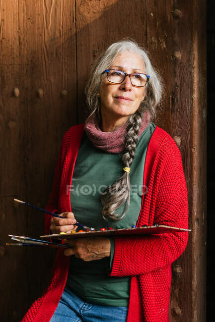 Encantada artista feminina sênior com paleta de tinta e pincéis de pé perto da porta de madeira e olhando para longe — Fotografia de Stock