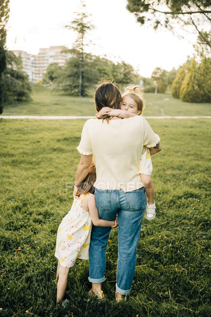 Rückansicht des niedlichen Mädchens und Blick in die Kamera, während Mutter mit kleiner Schwester an den Händen während des Sommertages zusammen im grünen Park kuschelt — Stockfoto