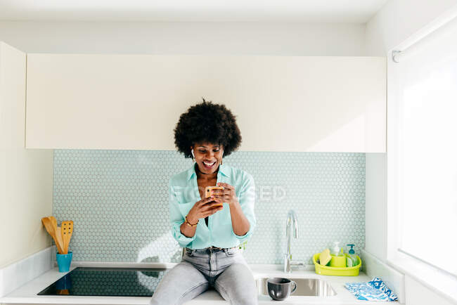 Joven mujer afroamericana positiva en ropa casual con auriculares inalámbricos escuchando música y charlando en el teléfono inteligente mientras está sentada en el mostrador de la cocina en casa - foto de stock