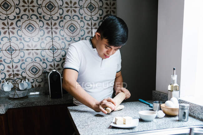 Enfoque adolescente étnico con síndrome de Down rodando masa con rodillo mientras cocina en la cocina - foto de stock