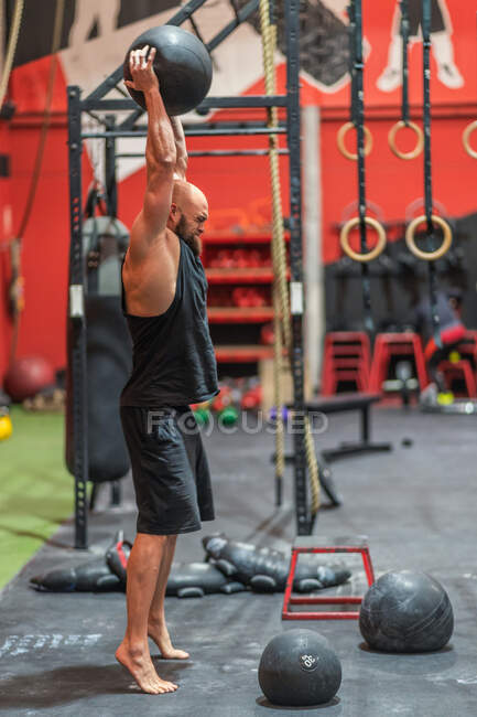 Full length side view of strong male athlete lifting heavy ball in raised arms while exercising in contemporary gym — Stock Photo