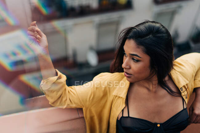 Von oben lehnt eine hispanische Frau im gelben Hemd an Absperrgitter und schaut weg, während sie es sich auf dem Balkon gemütlich macht — Stockfoto