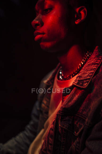Crop calm stylish African American man in jeans jacket under neon red light in shade on black background looking away — Stock Photo