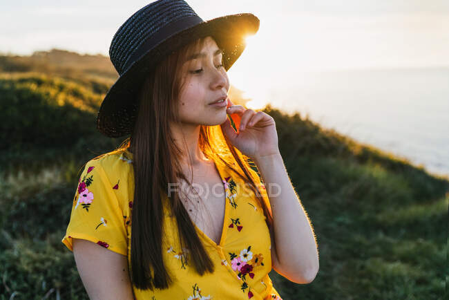 Attractive young female in yellow sundress and hat standing on verdant grassy meadow in sunny countryside — Stock Photo