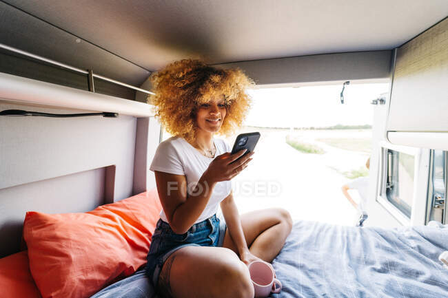 Anonymous woman with Afro hairstyle sitting upside upon bed in trailer and browsing mobile phone on sunny summer day — Stock Photo