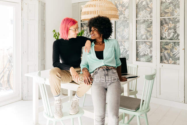 Веселі молоді стильні багаторасові друзі-жінки дивляться один на одного, сидячи разом на столі у світлій кімнаті — стокове фото