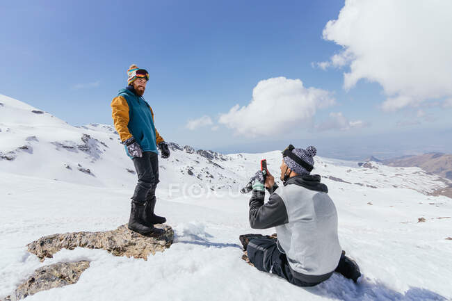 Мужчина в теплой одежде фотографирует веселого партнера по сотовому телефону на хребте под облачно-голубым небом зимой — стоковое фото
