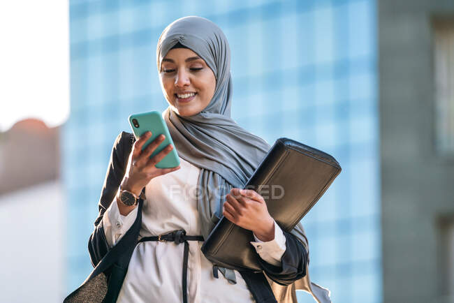 Счастливая мусульманка-предприниматель в хиджабе и с папкой, стоящей на улице просматривает на мобильном телефоне во время обсуждения бизнес-проекта — стоковое фото