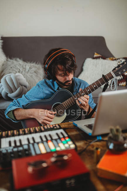 Jovem em fones de ouvido tocando na guitarra perto da mesa com laptop e sintetizador em casa olhando para a câmera — Fotografia de Stock