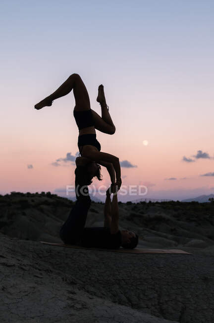 Vista lateral da mulher flexível equilibrando de cabeça para baixo enquanto pratica acroioga com parceiro masculino contra o céu do pôr-do-sol nas montanhas — Fotografia de Stock