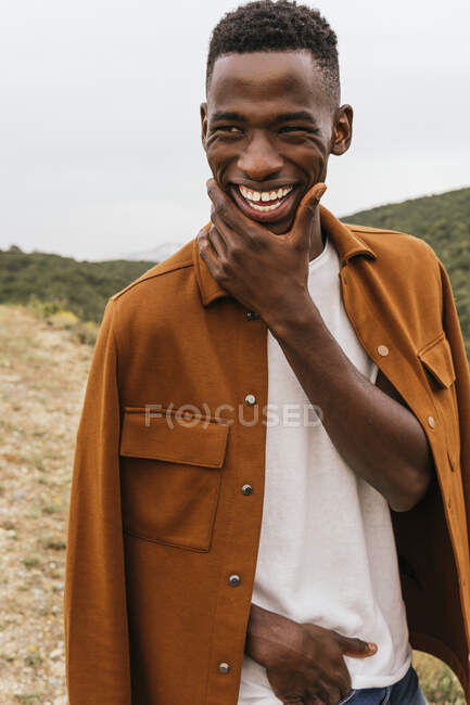 Modèle masculin afro-américain positif en tenue tendance debout avec la main dans la poche et le visage touchant dans la nature — Photo de stock