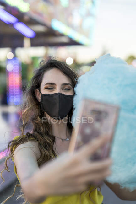 Женщина в защитной маске и с голубой сладкой сладкой сахарной ватой делает самоснимок на мобильном телефоне, веселясь на ярмарке — стоковое фото
