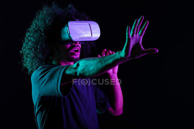 Волнующий латинский мужчина с прической афро и в очках виртуальной реальности на черном фоне в студии с неоновыми огнями — стоковое фото
