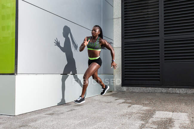 Vista lateral de la atleta afroamericana saltando hacia adelante mientras corre cerca de la pared del edificio moderno en la calle de la ciudad - foto de stock
