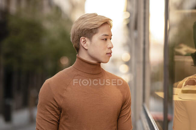 Vista lateral do modelo masculino asiático bonito com cabelo loiro olhando para a câmera na rua da cidade — Fotografia de Stock