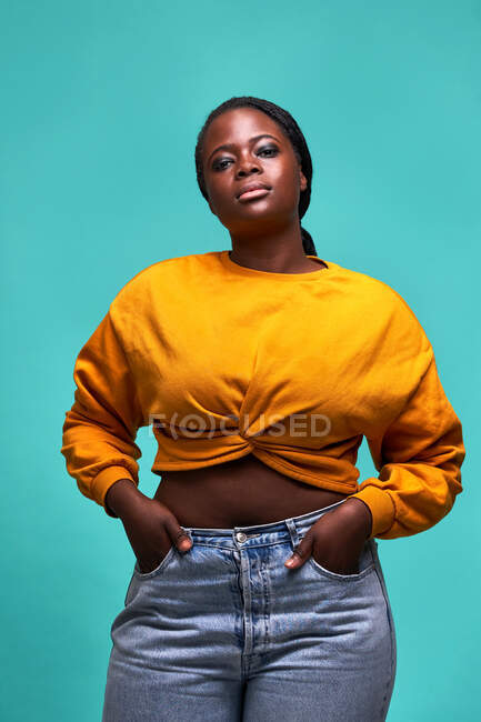Unemotional grassoccio afroamericano femminile in maglione giallo in piedi guardando la fotocamera con le mani in tasca contro il muro blu — Foto stock