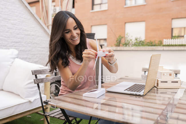 Encantada arquitecta montando el modelo plástico del molino de viento mientras está sentada en la mesa con el portátil y trabajando en el proyecto en la terraza - foto de stock