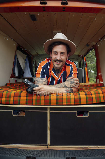Lächelnder Reisender mit Fotokamera, der im Van auf dem Bett liegt und in die Kamera schaut, während er den Sommerurlaub genießt — Stockfoto