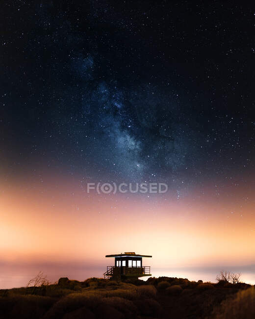 Espectacular paisaje de cielo estrellado con la Vía Láctea brillando sobre la torre de salvavidas ubicada en la colina por la noche - foto de stock