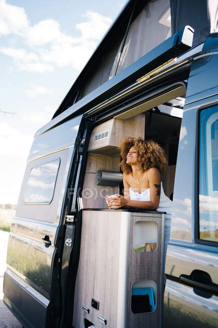 Angolo basso di rilassato afro-americano femminile con tazza di bevanda calda occhi di chiusura e refrigerante all'interno moderno camper in estate mattina — Foto stock