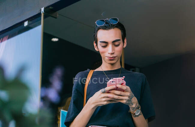 Bajo ángulo de mensajería masculina gay en las redes sociales en el teléfono móvil mientras está de pie en la calle y sonriendo - foto de stock