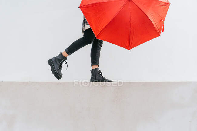 Jovem anônima com guarda-chuva vermelho andando e equilibrando na fronteira contra a parede cinza no dia chuvoso na rua — Fotografia de Stock