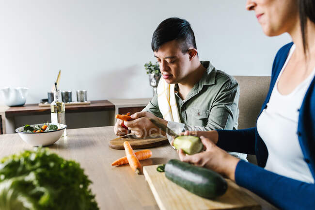 Cropped anonymous mother and teenage son with Down syndrome sitting at table and cutting vegetables while preparing salad for lunch at home — Stock Photo