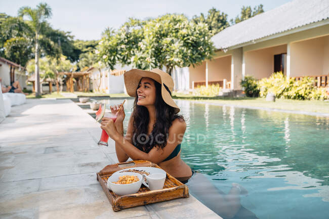 Cheerful female tourist in swimwear enjoying refreshing drink while leaning on poolside with tasty breakfast on tray in tropical resort — Stock Photo