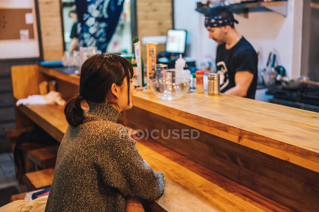 Vista lateral de la mujer asiática en ropa casual sentado en el mostrador y hablando con el trabajador masculino del moderno bar ramen - foto de stock
