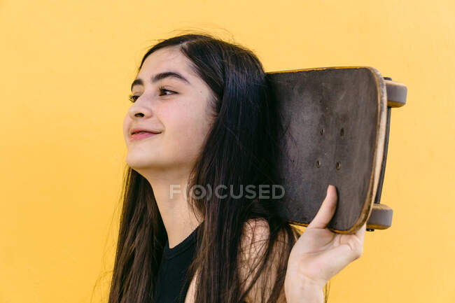 Vue latérale de joyeux jeune skateboarder féminin tenant skateboard derrière la tête tout en regardant loin — Photo de stock