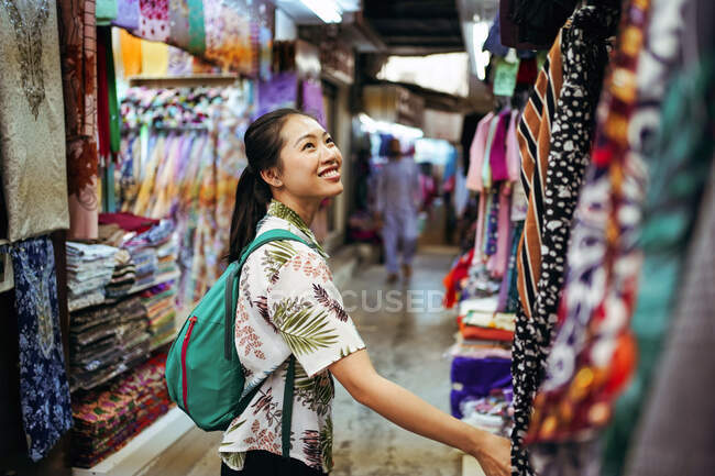 Side view of positive young female traveler in casual clothes with backpack standing near stand with colorful fabric while visiting Mutrah Souq marketplace in Muscat in Oman — Stock Photo