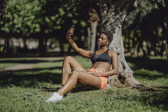 African American woman in sportswear holding smartphone and taking a selfie while sitting on grass in sunny day — Stock Photo