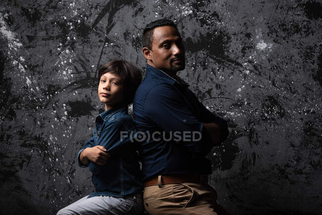 Père multiracial et fils adolescent assis dos à dos avec les bras croisés sur fond sombre en studio et regardant la caméra — Photo de stock