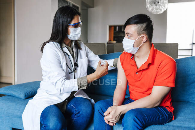 Médico feminino com seringa que faz a injeção da vacina para o menino adolescente latino com síndrome de Down em casa durante o coronavírus — Fotografia de Stock