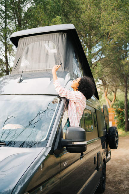 Side view of young African American female traveler leaning out open window of camper van and taking selfie on smartphone while enjoying summer journey in forest — Stock Photo