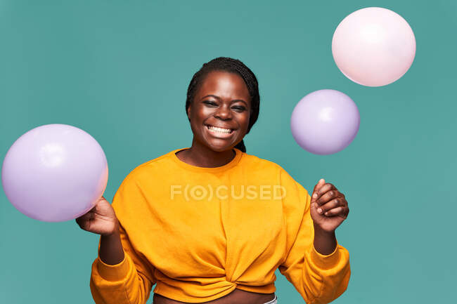Mulher afro-americana excitada em jeans e top amarelo perto de cair balões contra fundo azul em estúdio — Fotografia de Stock