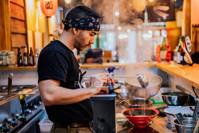 Seitenansicht eines Mannes mit Kopftuch, der am Tresen steht und in einem modernen asiatischen Café Ramen kocht — Stockfoto