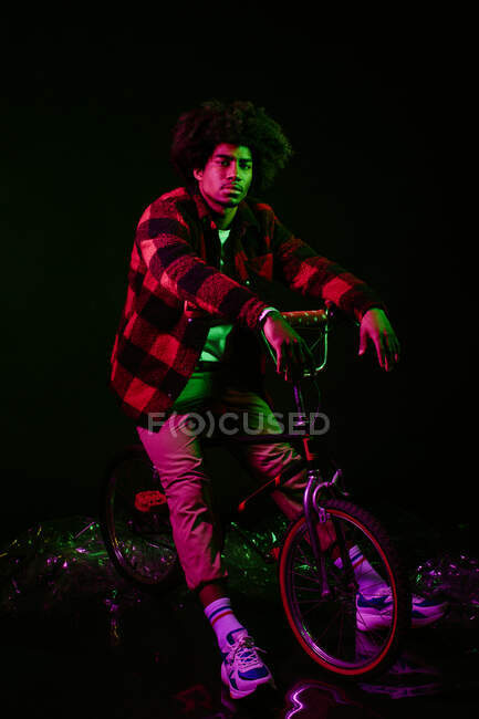 Confiant jeune homme afro-américain aux cheveux bouclés en tenue tendance équitation vélo rétro sur fond violet — Photo de stock