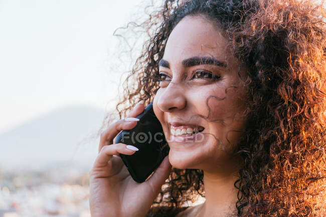 Agréable jeune femme hispanique avec de longs cheveux bouclés parlant sur téléphone portable et souriant joyeusement le soir ensoleillé d'été en plein air — Photo de stock