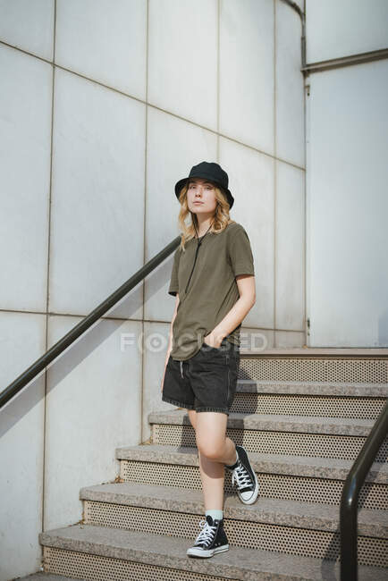 Молодая женщина в повседневной одежде смотрит на камеру, стоящую на лестнице напротив бетонной стены современного здания на городской улице днем — стоковое фото